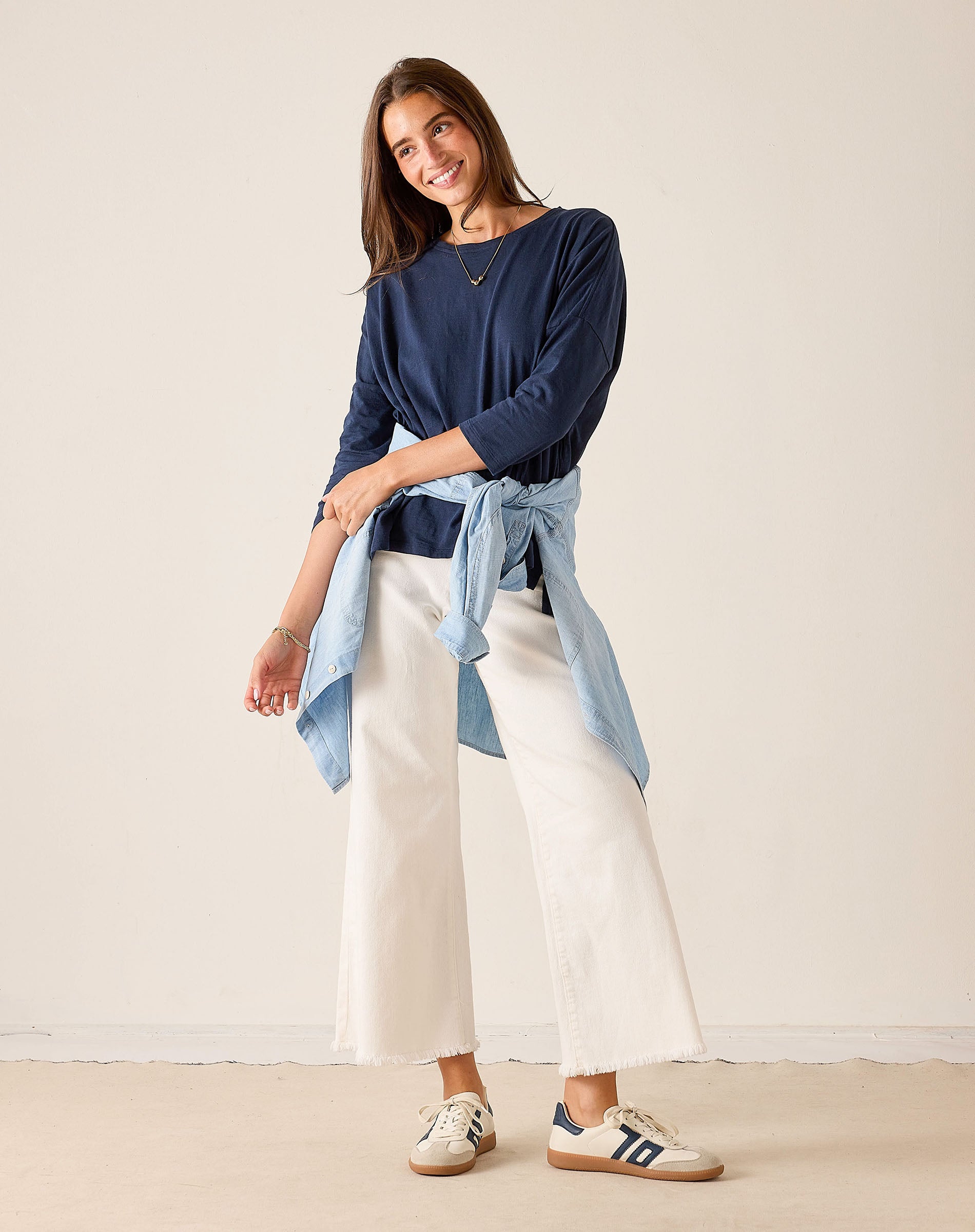 Woman wearing a navy top and white pants with a light blue shirt tied around her waist against a plain background