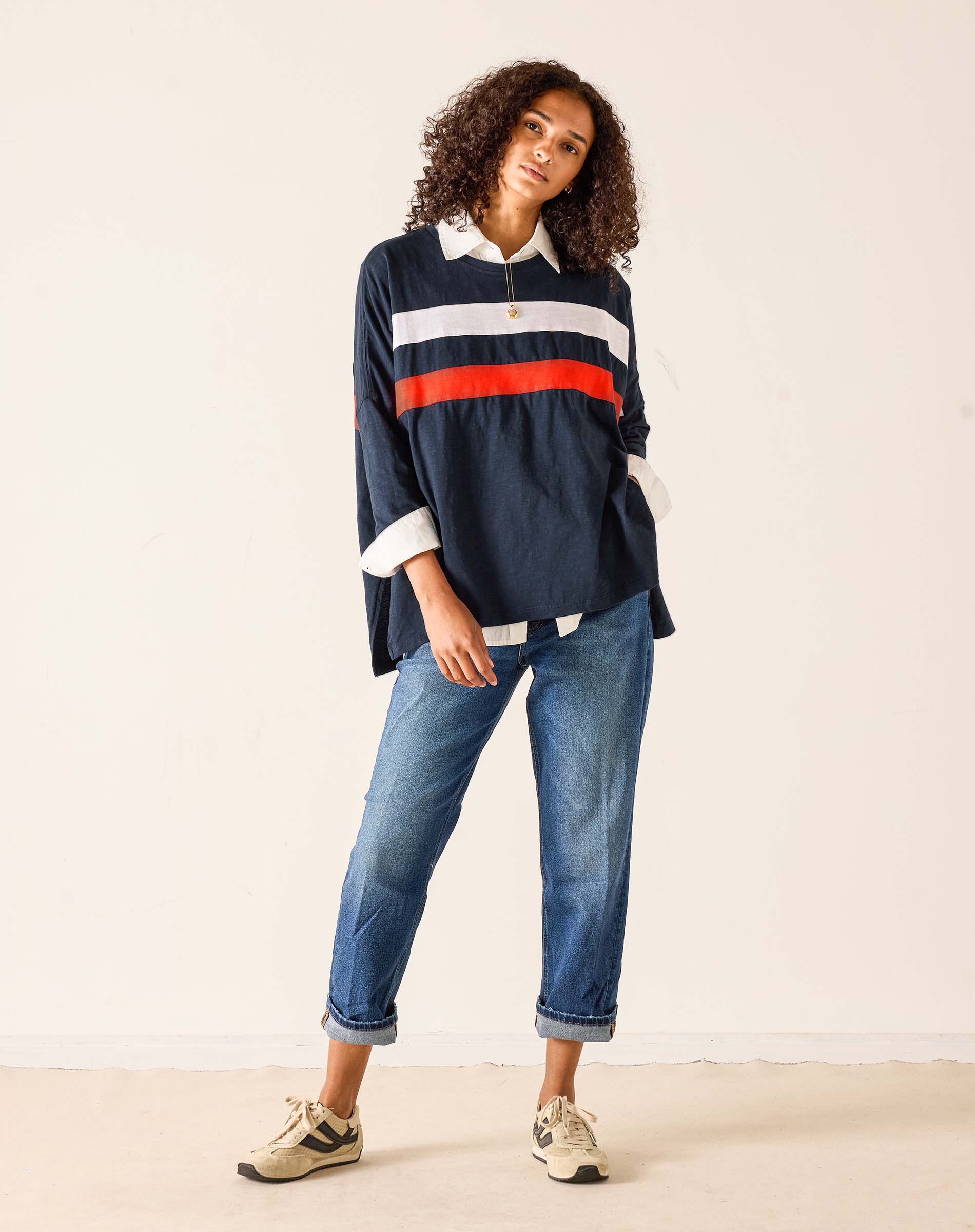 Woman wearing a navy sweater with red and white stripes and blue jeans on a plain background