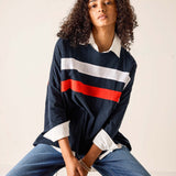 Woman wearing a navy rugby shirt with red and white stripes and blue jeans, sitting on a stool.