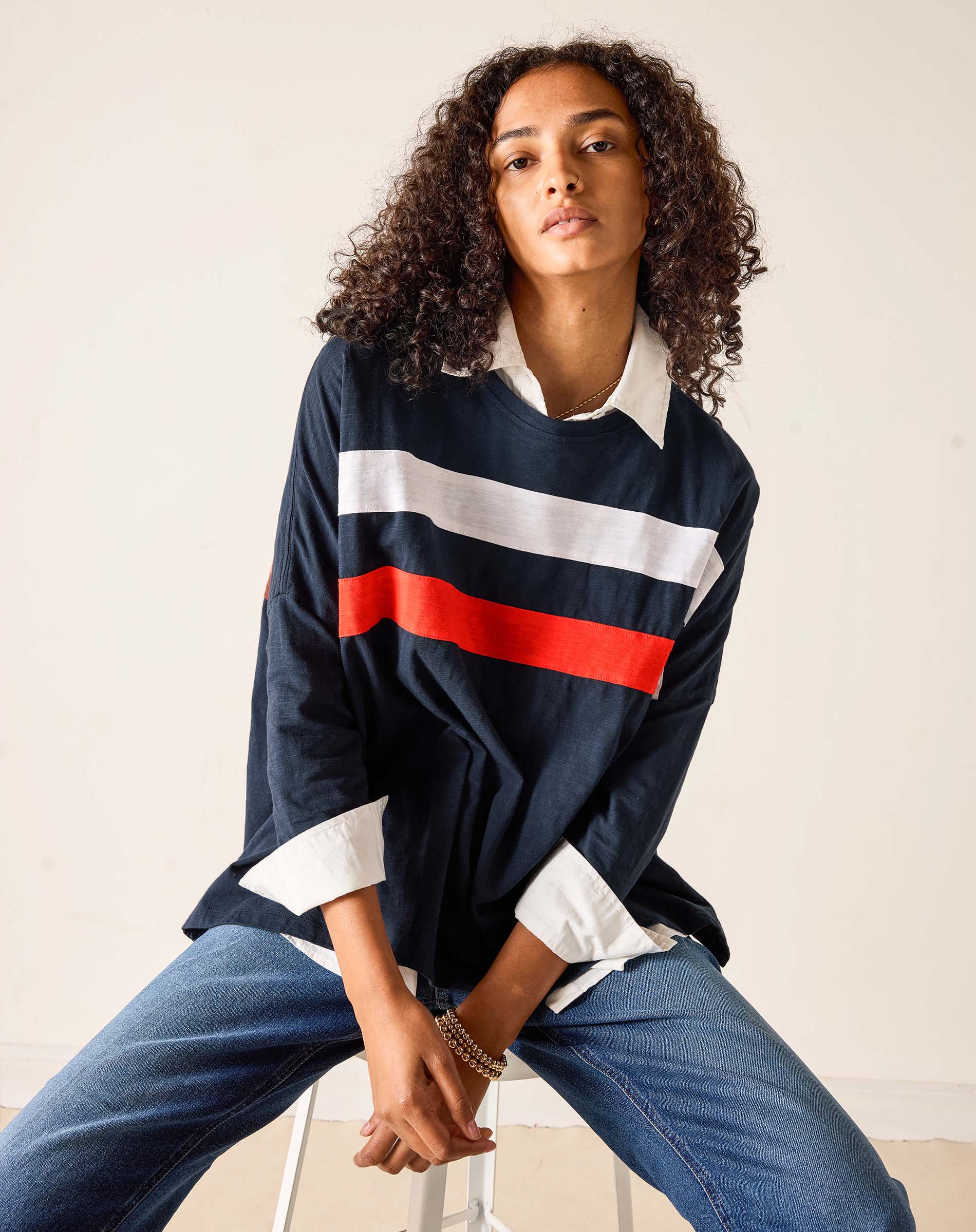 Woman wearing a navy rugby shirt with red and white stripes and blue jeans, sitting on a stool.
