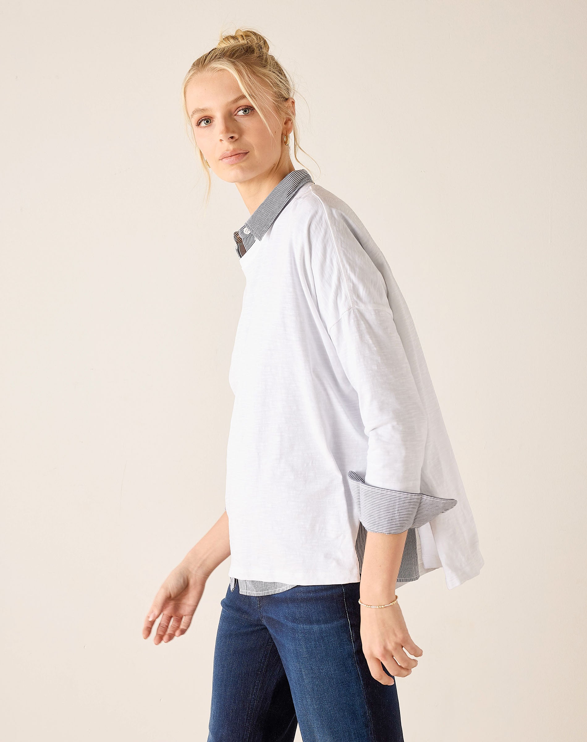 Woman wearing a white shirt with a blue collar and dark jeans on a plain background