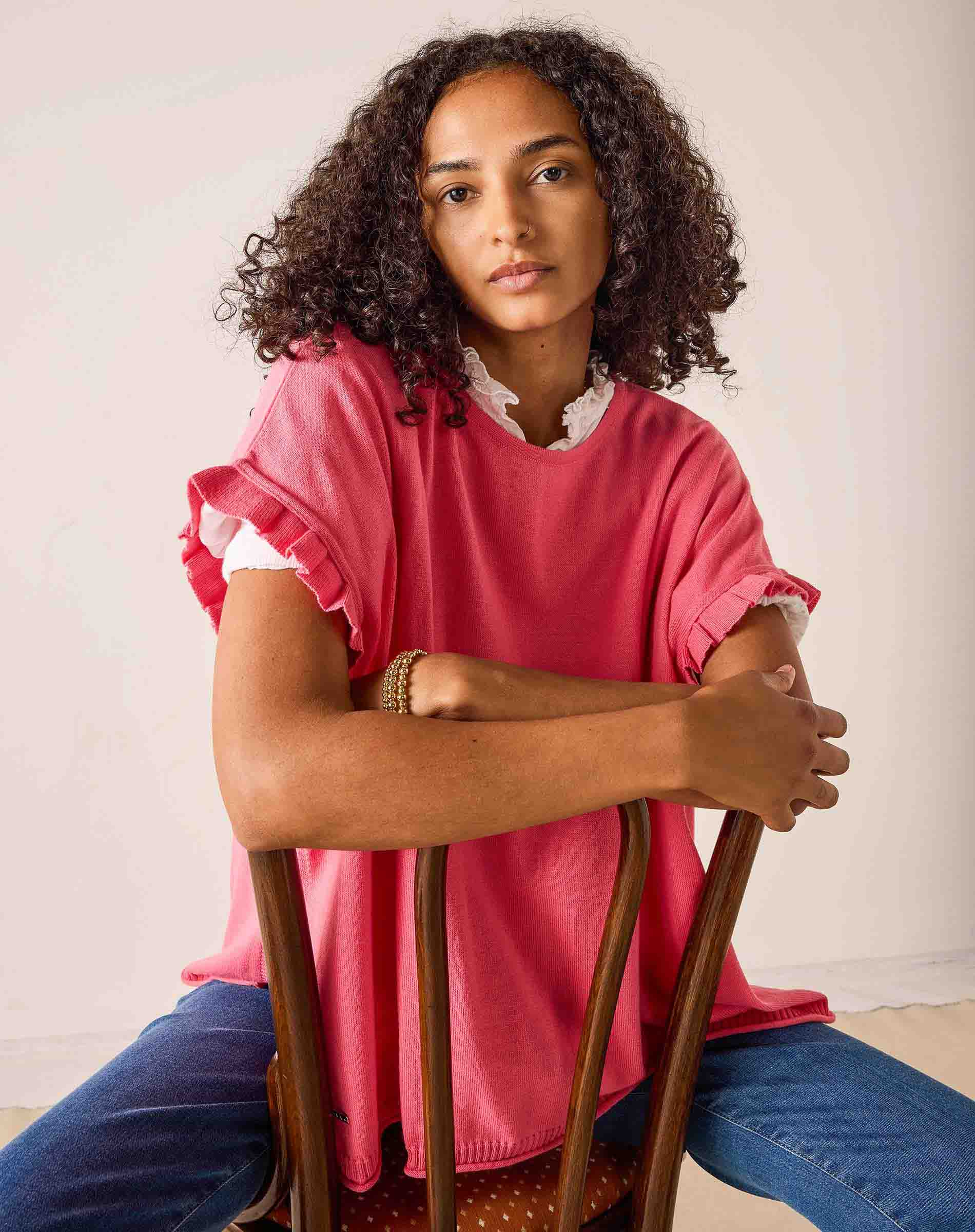 Woman sitting on a wooden chair wearing a pink top and blue jeans against a plain background