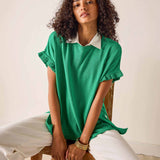 Woman wearing a green top with white collar and cuffs, sitting on a wooden chair against a white wall.