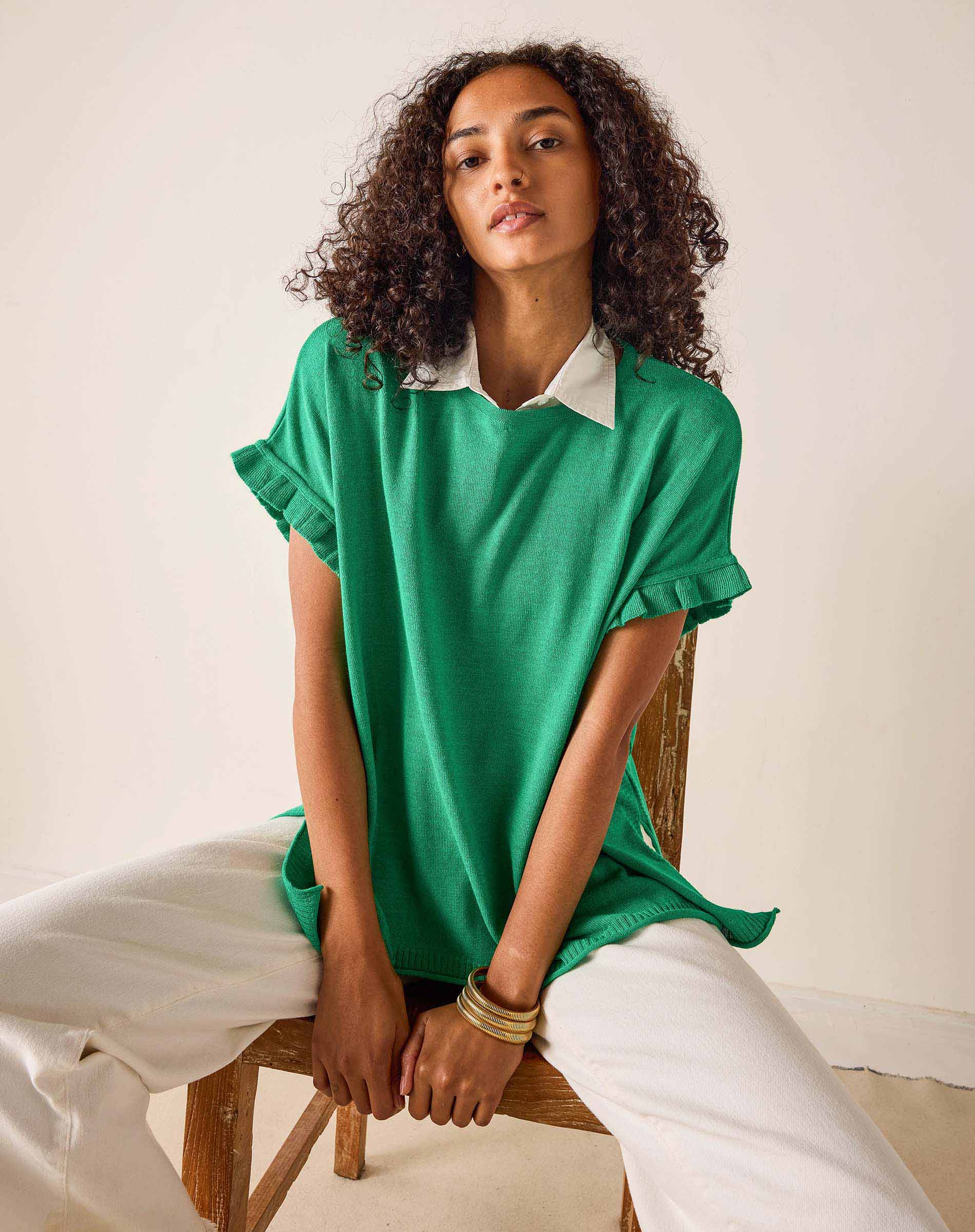 Woman wearing a green top with white collar and cuffs, sitting on a wooden chair against a white wall.