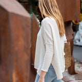 Woman wearing a white textured sweater and blue jeans standing outdoors with rusted metal sculptures in the background.