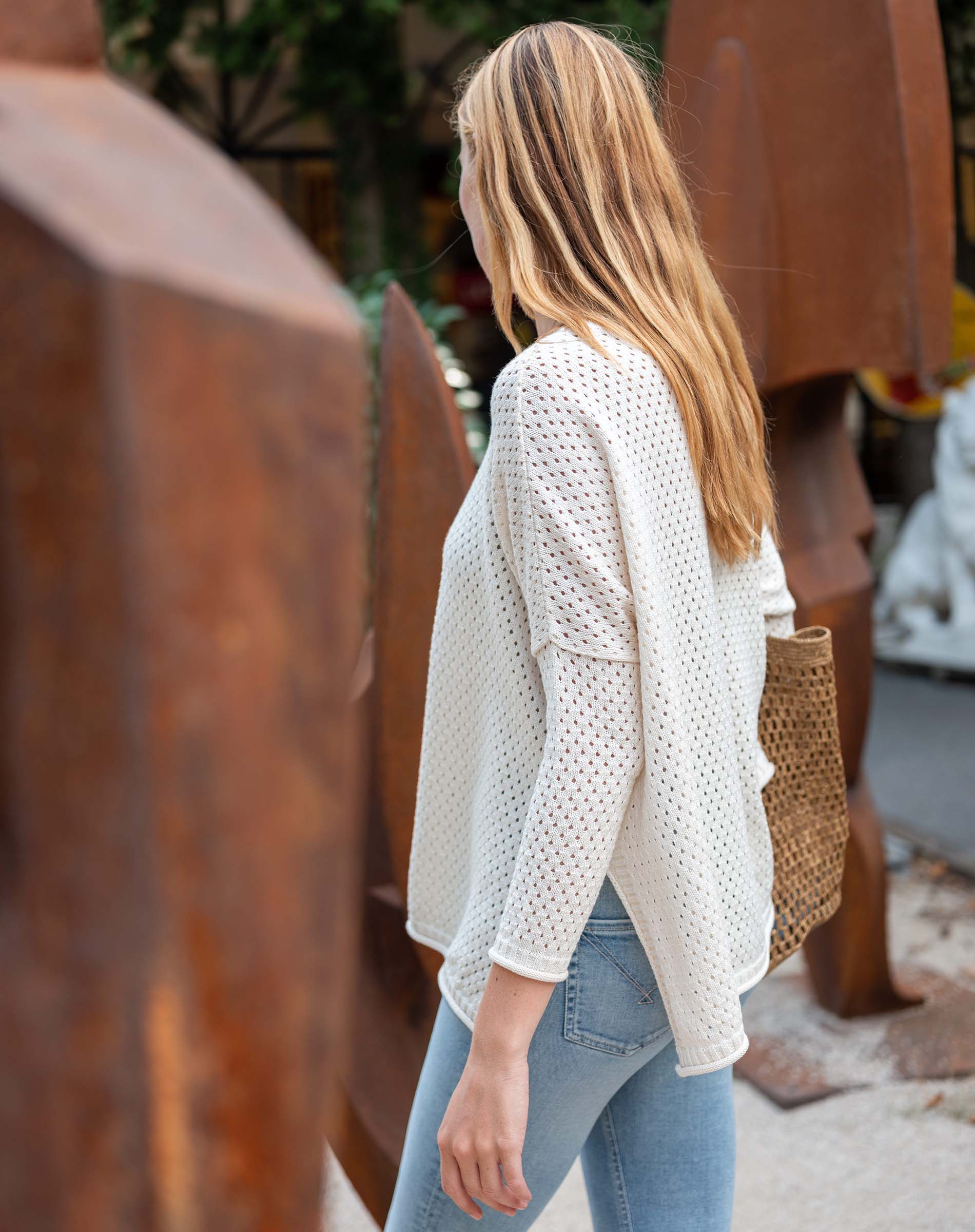 Woman wearing a white textured sweater and blue jeans standing outdoors with rusted metal sculptures in the background.