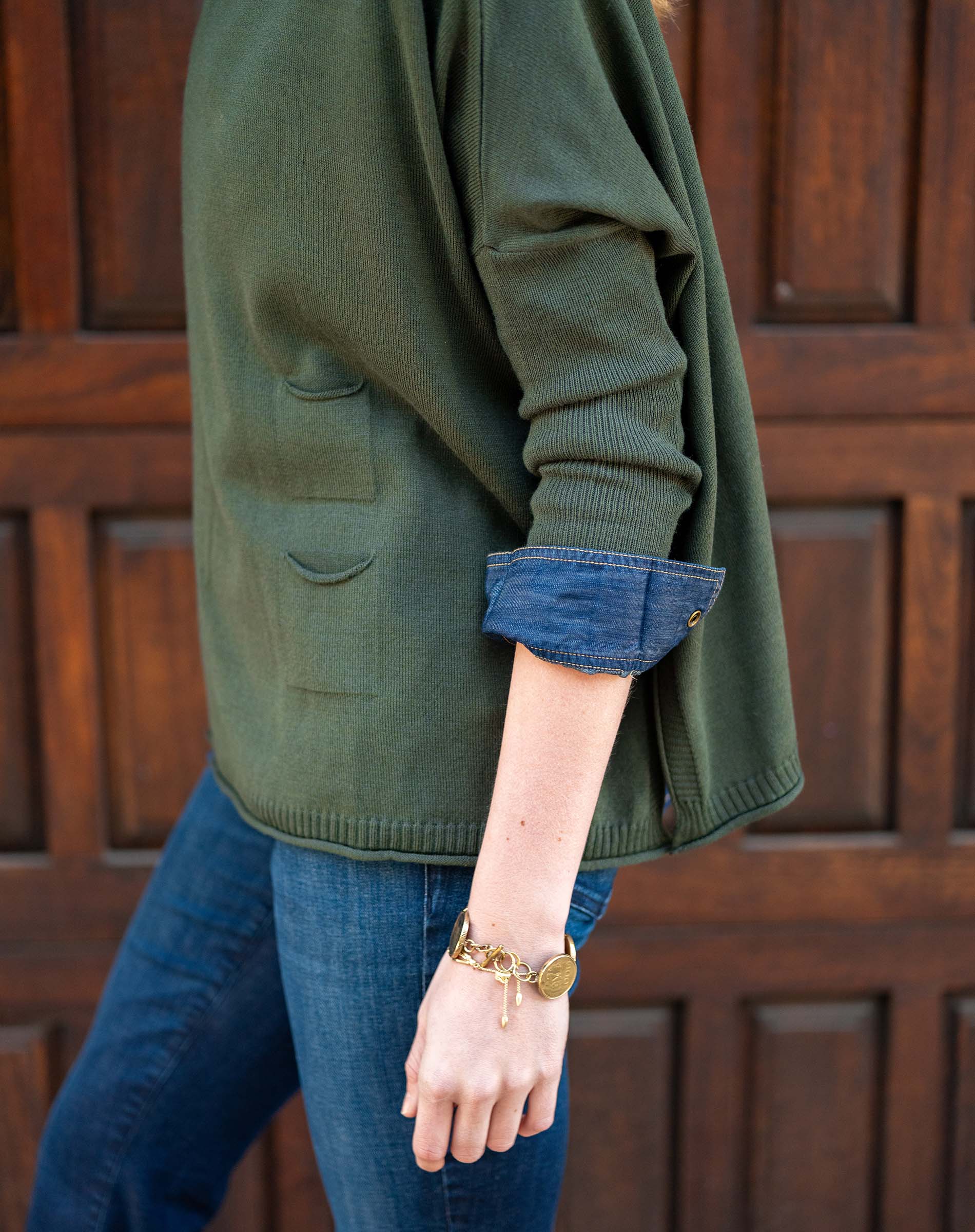 Person wearing a green sweater with rolled-up sleeves and blue jeans, standing against a wooden door.