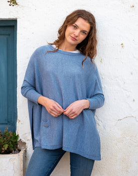 female wearing deep blue sweater over blue jeans leaning on a white wall