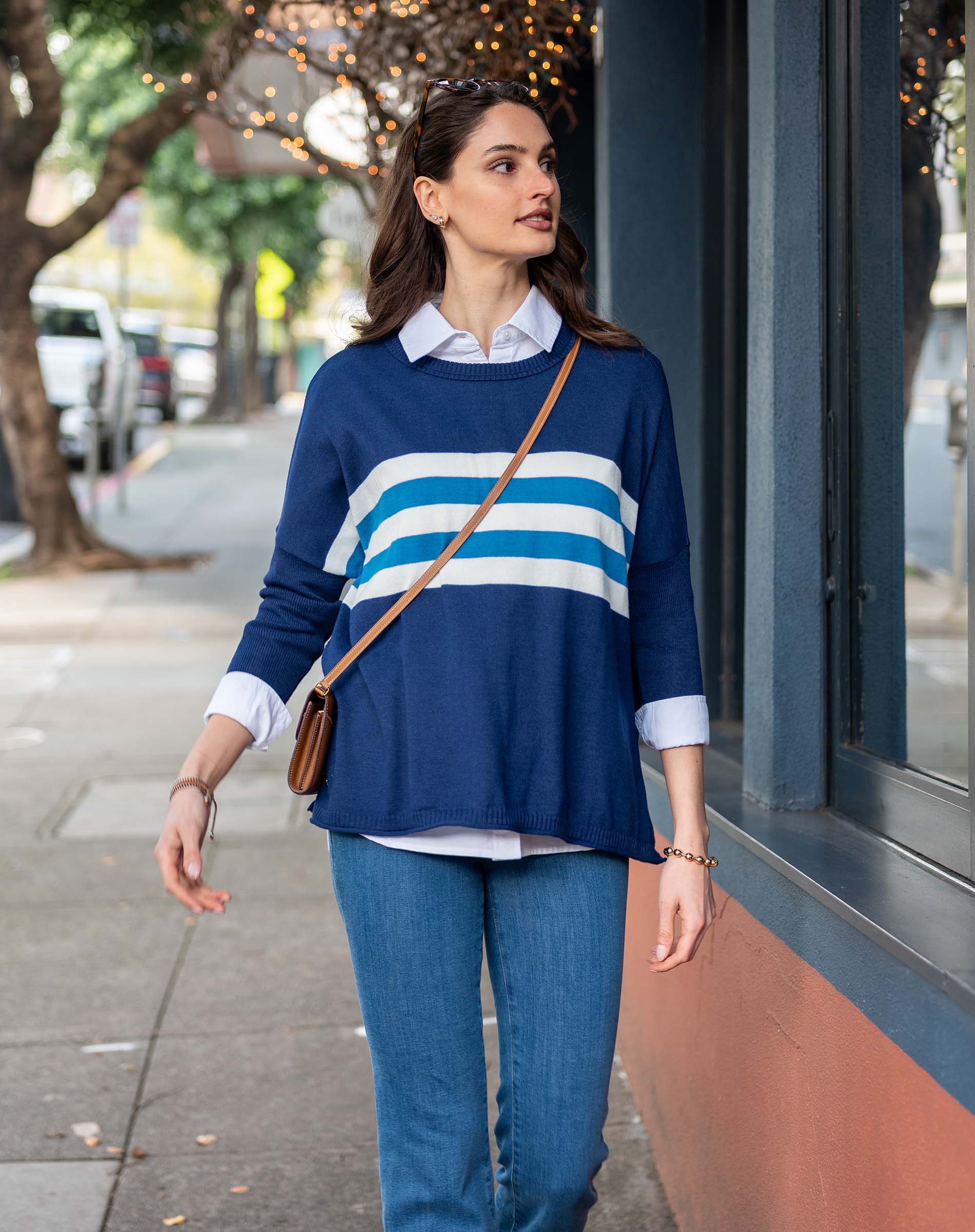 Woman wearing a blue sweater with white stripes and blue jeans, standing on a sidewalk.