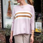 Woman wearing a light pink sweater with brown and white stripes, beige pants, and a beige cap outdoors.