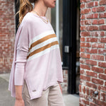 Woman wearing a light pink sweater with gold stripes and beige pants, standing against a brick wall.
