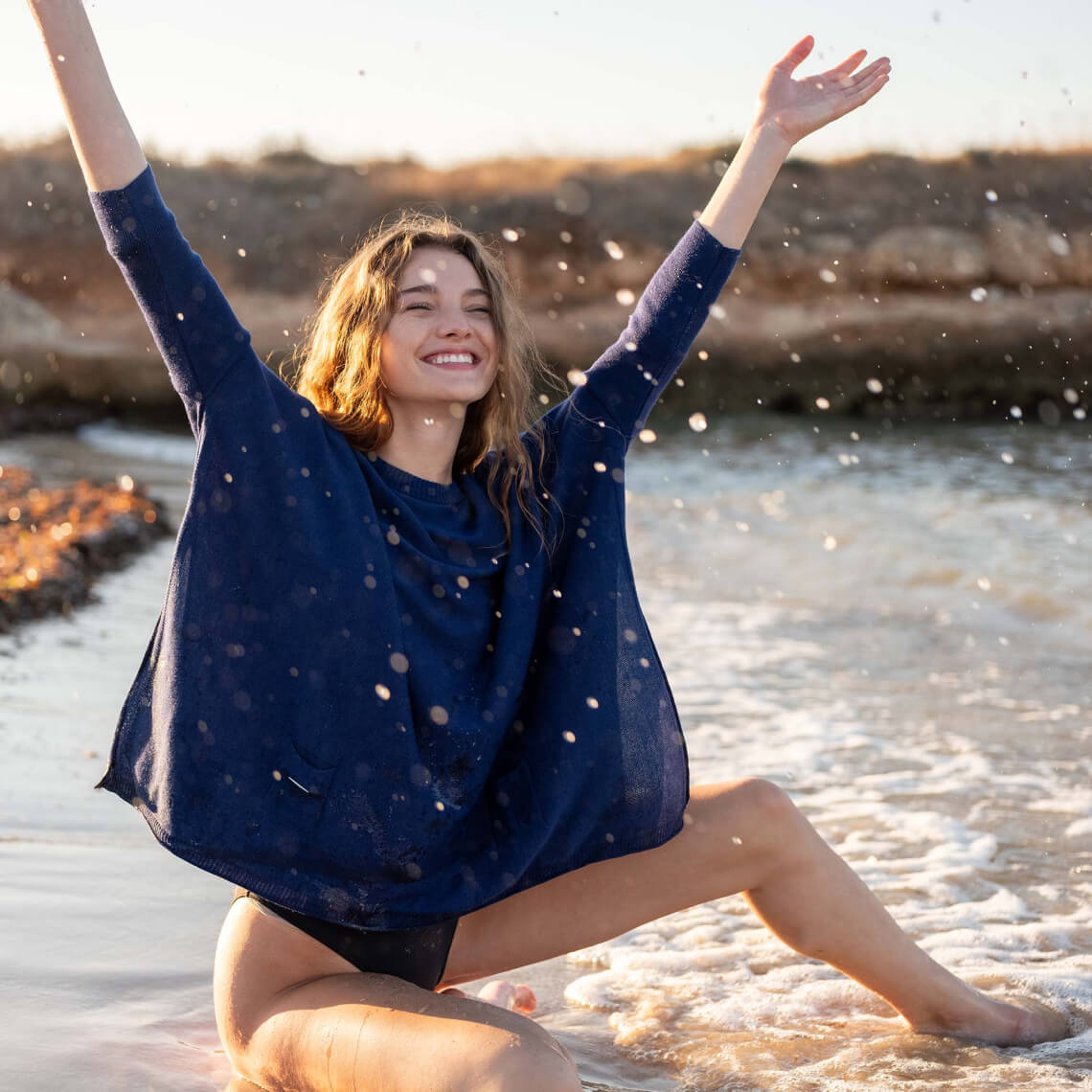 women wearing deepwater catalina sweater throwing water in the air sitting on the beach in a swimsuit