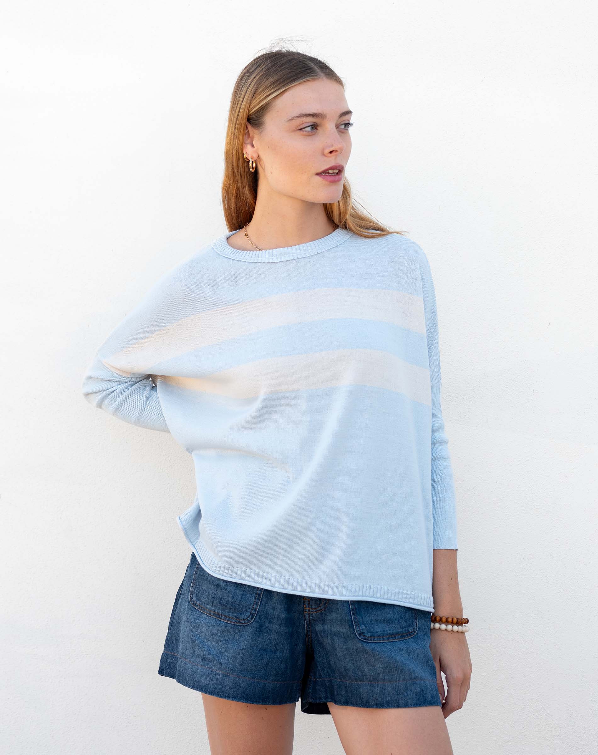 Woman wearing a light blue sweater with a white stripe and denim shorts against a white background