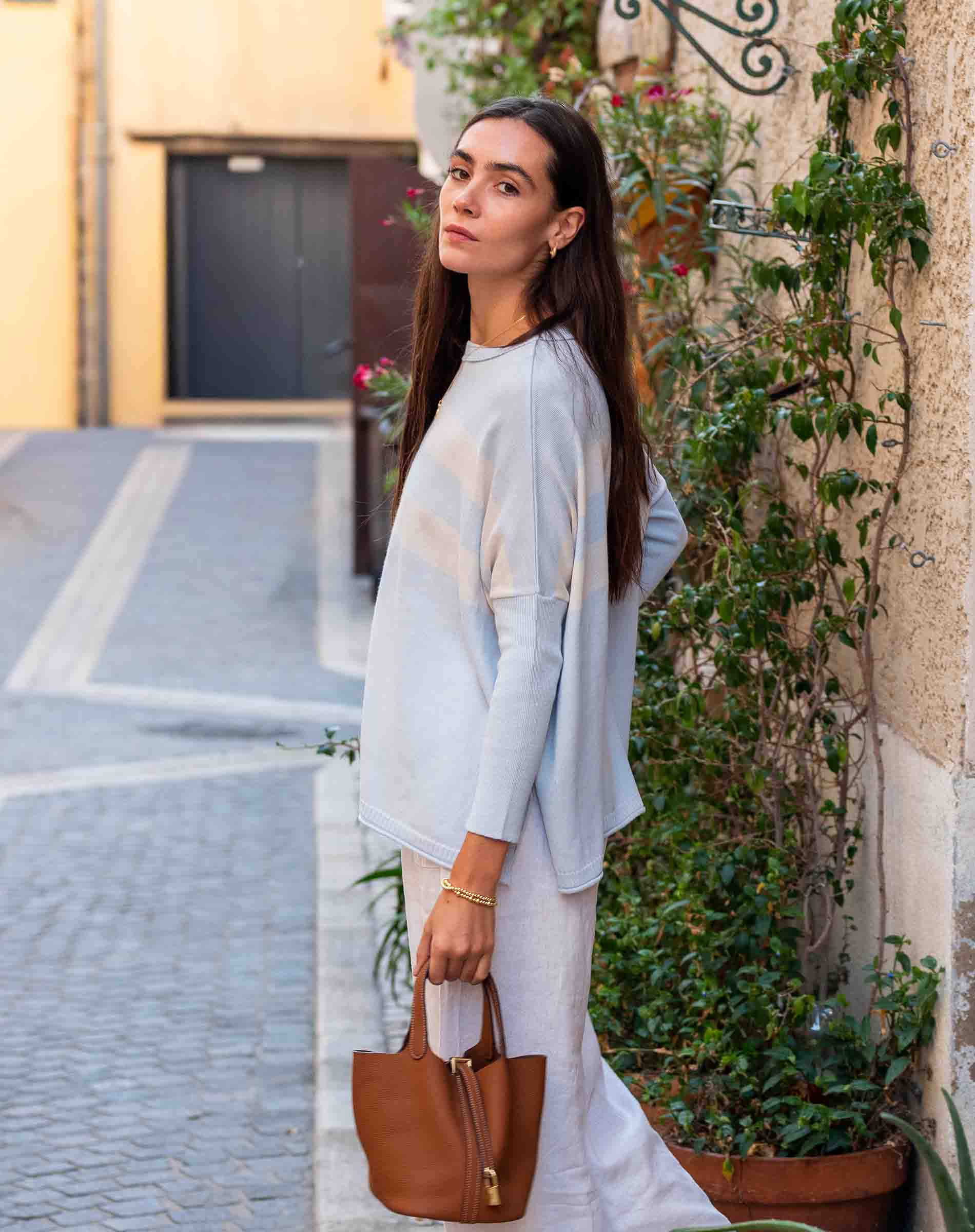 Woman in light blue top and white pants holding a brown bag on a street.