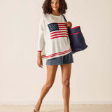 Woman wearing a sweater with an American flag design, holding a tote bag, on a plain background
