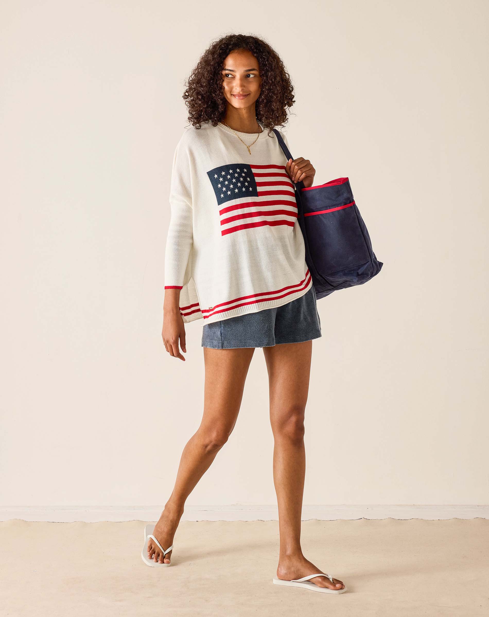 Woman wearing a sweater with an American flag design, holding a tote bag, on a plain background