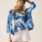 Person wearing a blue tie-dye shirt and white shorts with a cap on a plain background