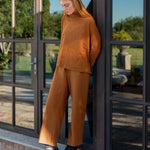 Person wearing a brown outfit standing by a glass door with a garden view.