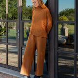 Person wearing a brown outfit standing by a glass door with a garden view.