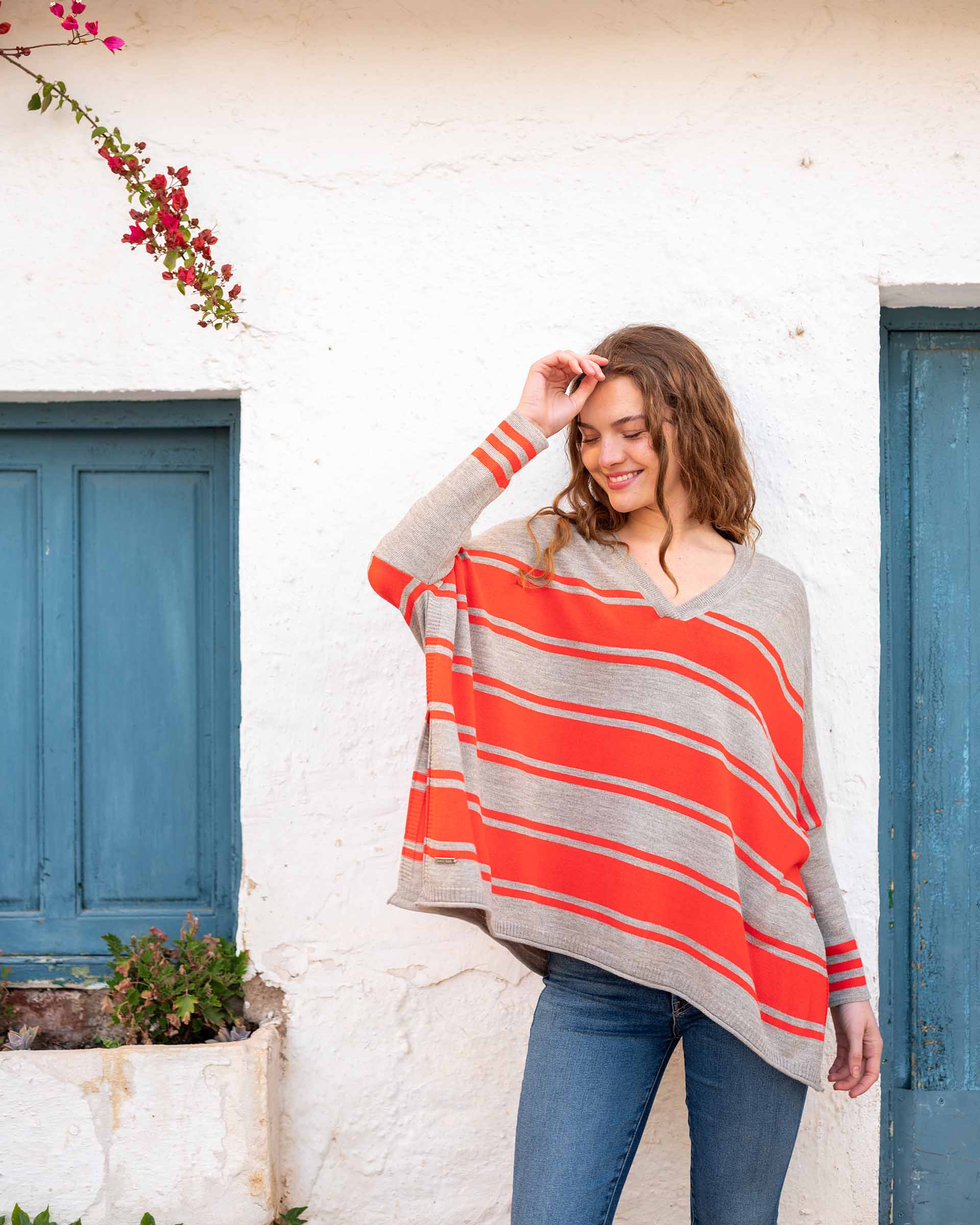 female wearing grey and red striped v neck sweater with blue jeans in front of white wall