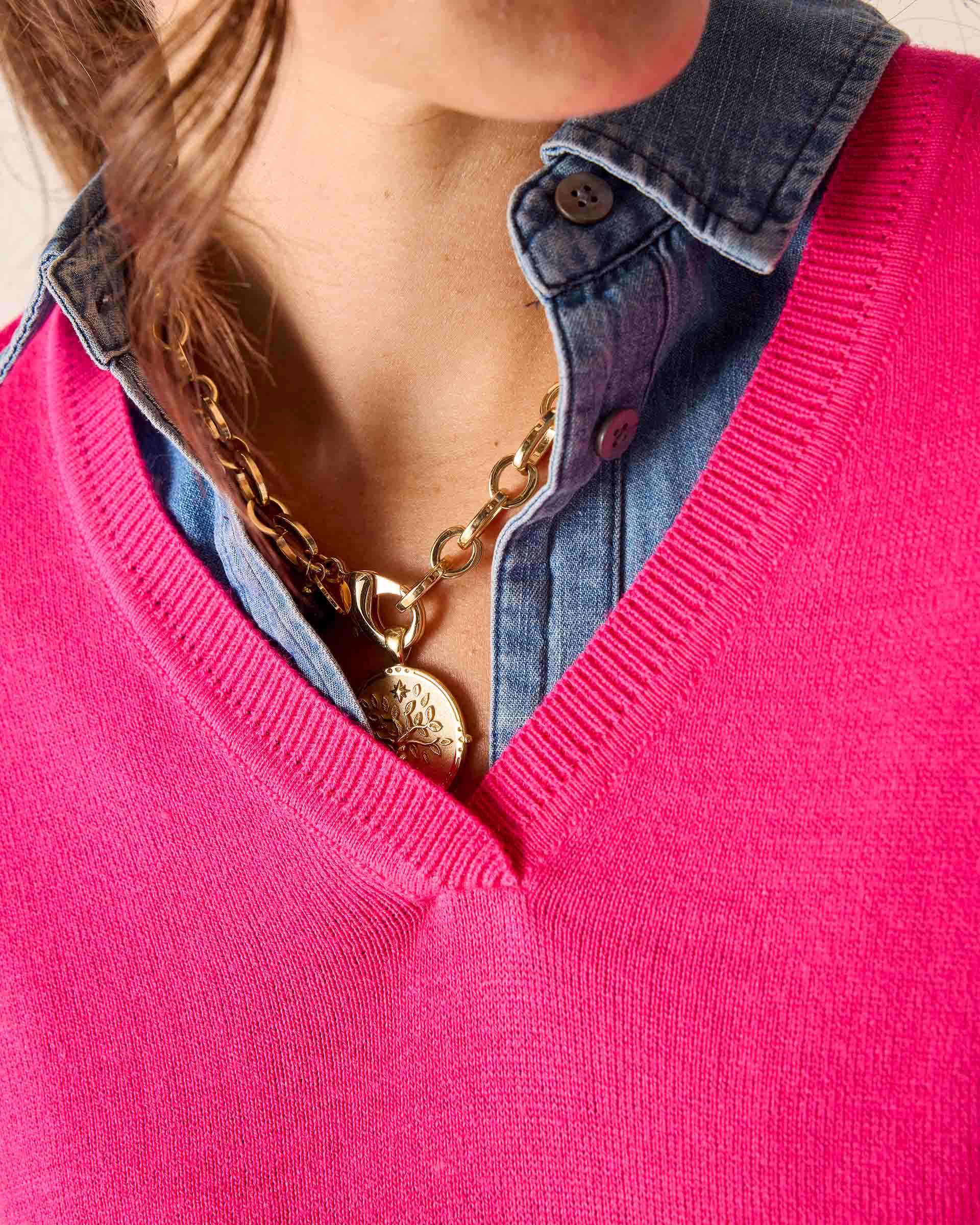Person wearing a pink sweater over a denim shirt with gold jewelry.