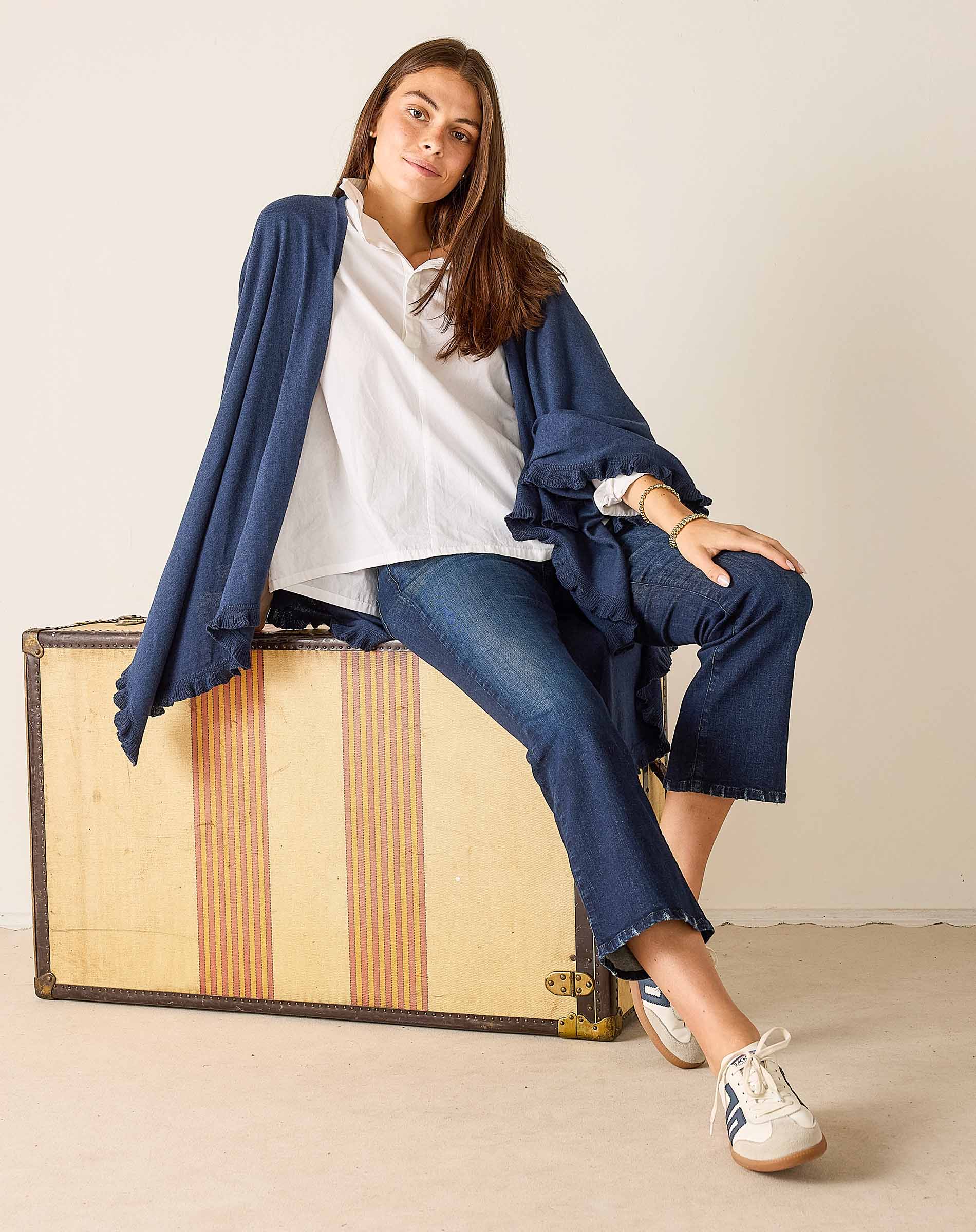 Woman sitting on a vintage suitcase wearing a blue jacket, white shirt, and jeans.