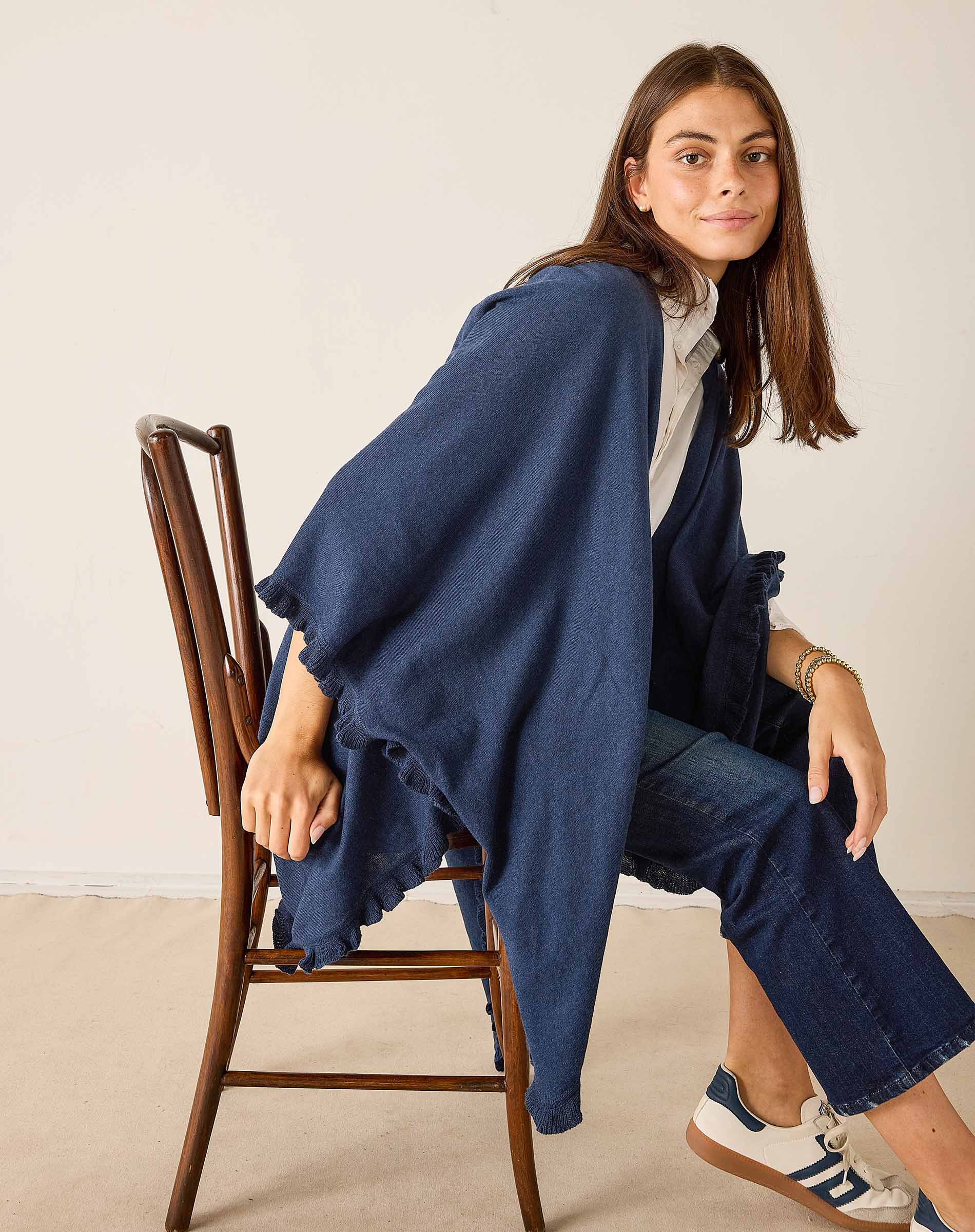 Woman sitting on a wooden chair wearing a blue poncho and white sneakers.