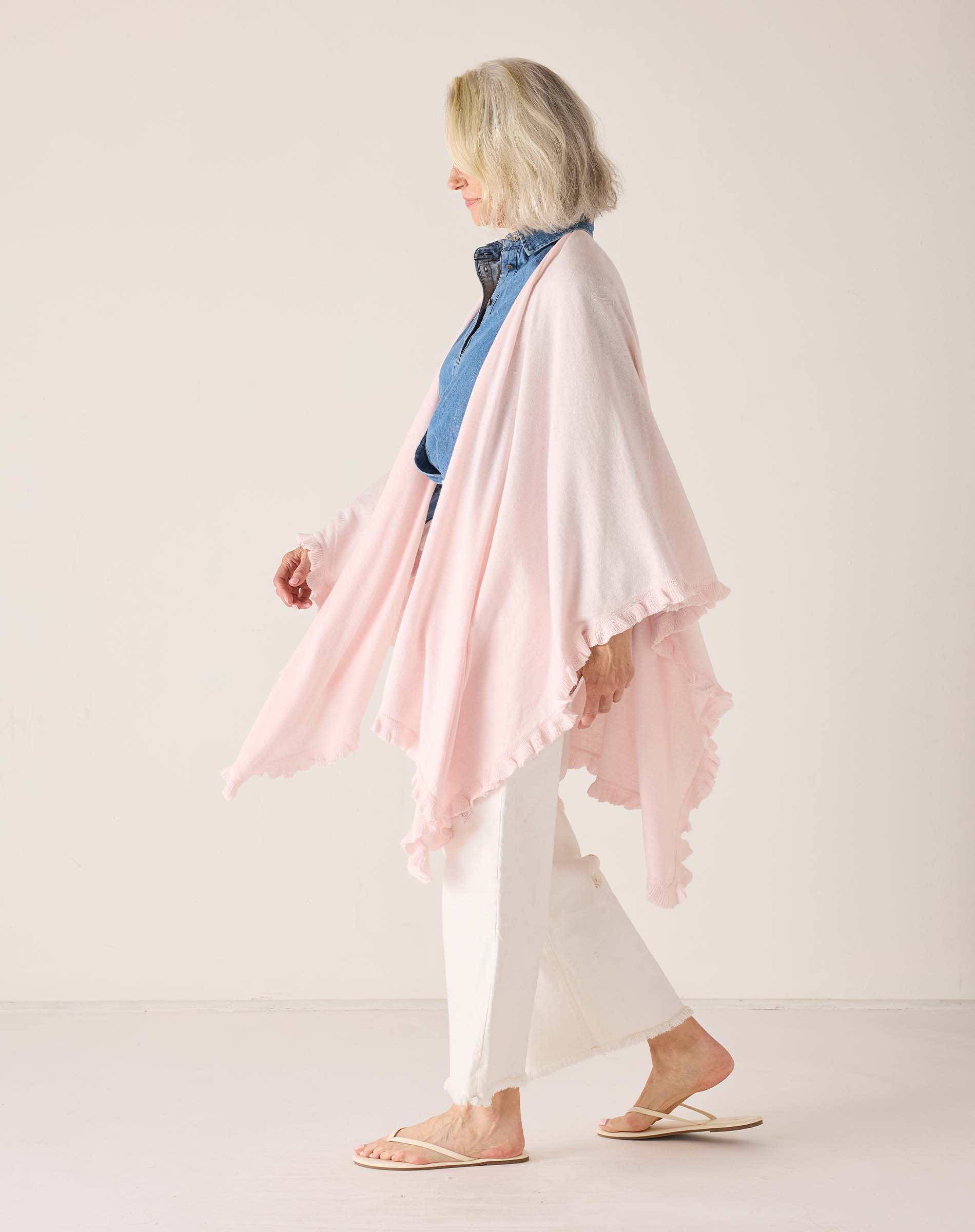 Woman wearing a light pink scarf over a blue top and white pants on a plain background