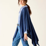 Woman wearing a blue cardigan over a patterned shirt and jeans on a plain background