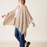 Woman wearing a beige poncho over a white shirt and blue jeans on a plain background