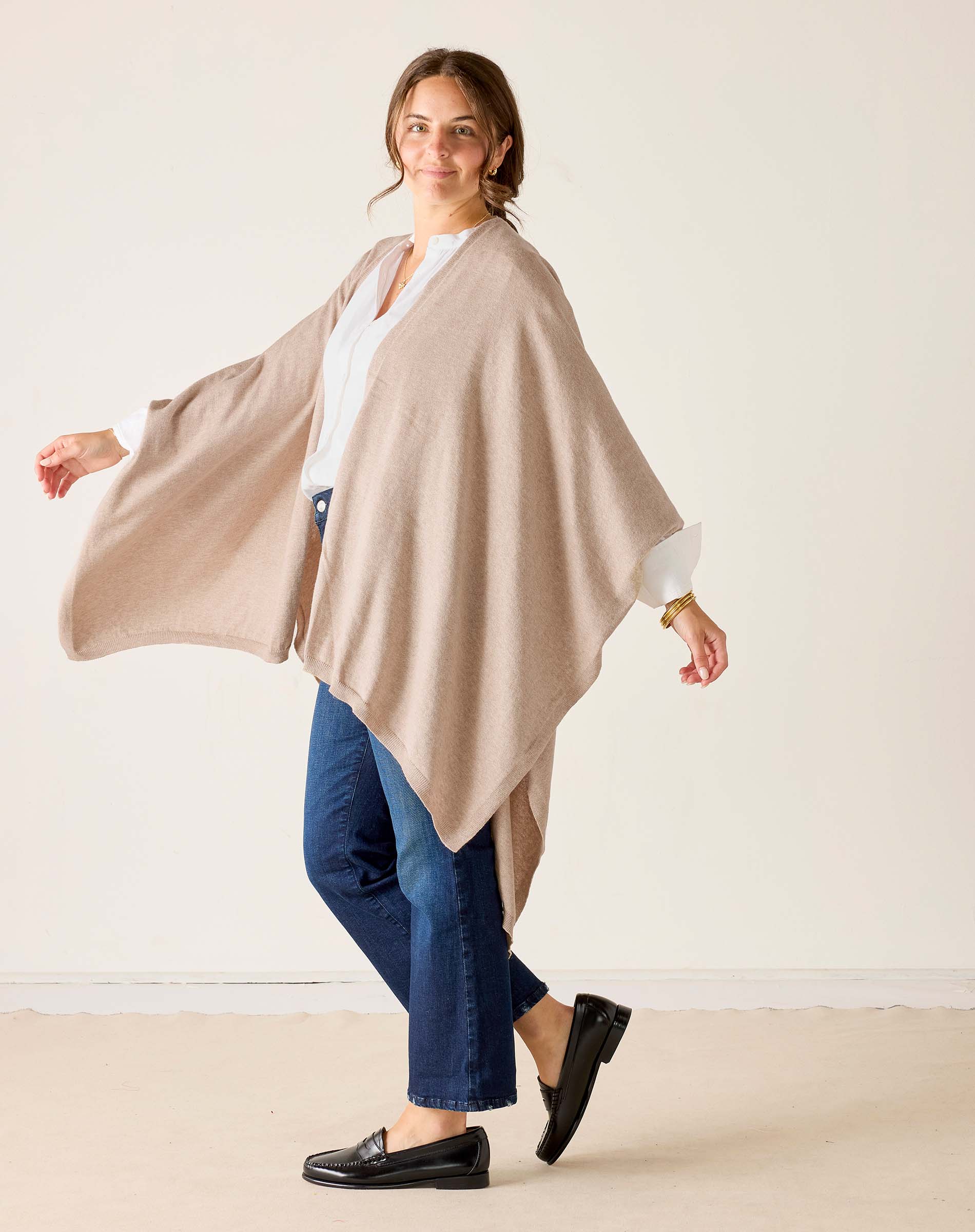 Woman wearing a beige poncho over a white shirt and blue jeans on a plain background