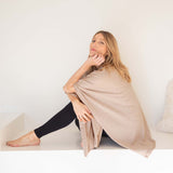 Woman sitting on a white surface with a beige blanket draped over her, against a plain white background