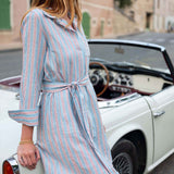 Woman in a striped dress standing next to a vintage car on a street.