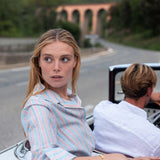Woman sitting in a convertible car with a man, looking surprised or confused, with a scenic background.