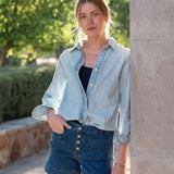Woman wearing a light blue denim jacket and shorts standing against a stone wall with greenery in the background.