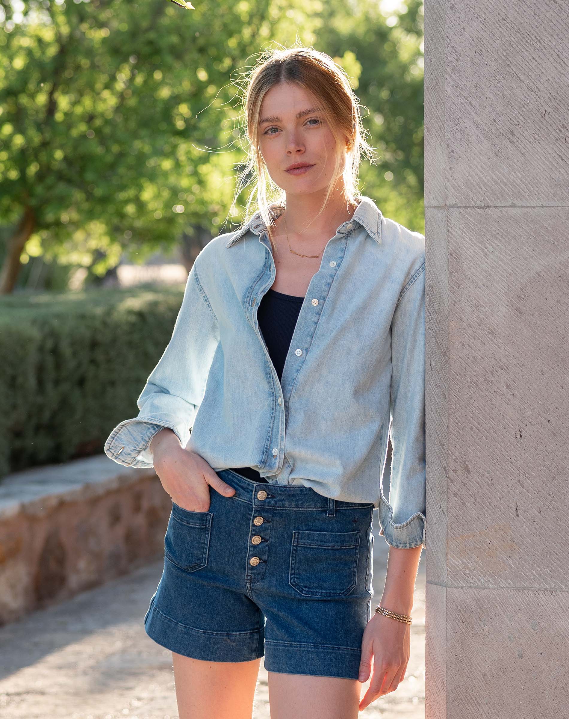 Woman wearing a light blue denim jacket and shorts standing against a stone wall with greenery in the background.