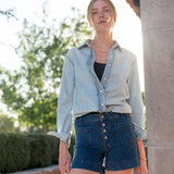 Woman wearing a light blue denim shirt and dark blue shorts standing outdoors.