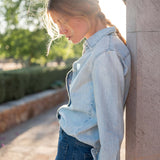 Woman wearing a light blue denim jacket and jeans leaning against a wall outdoors.