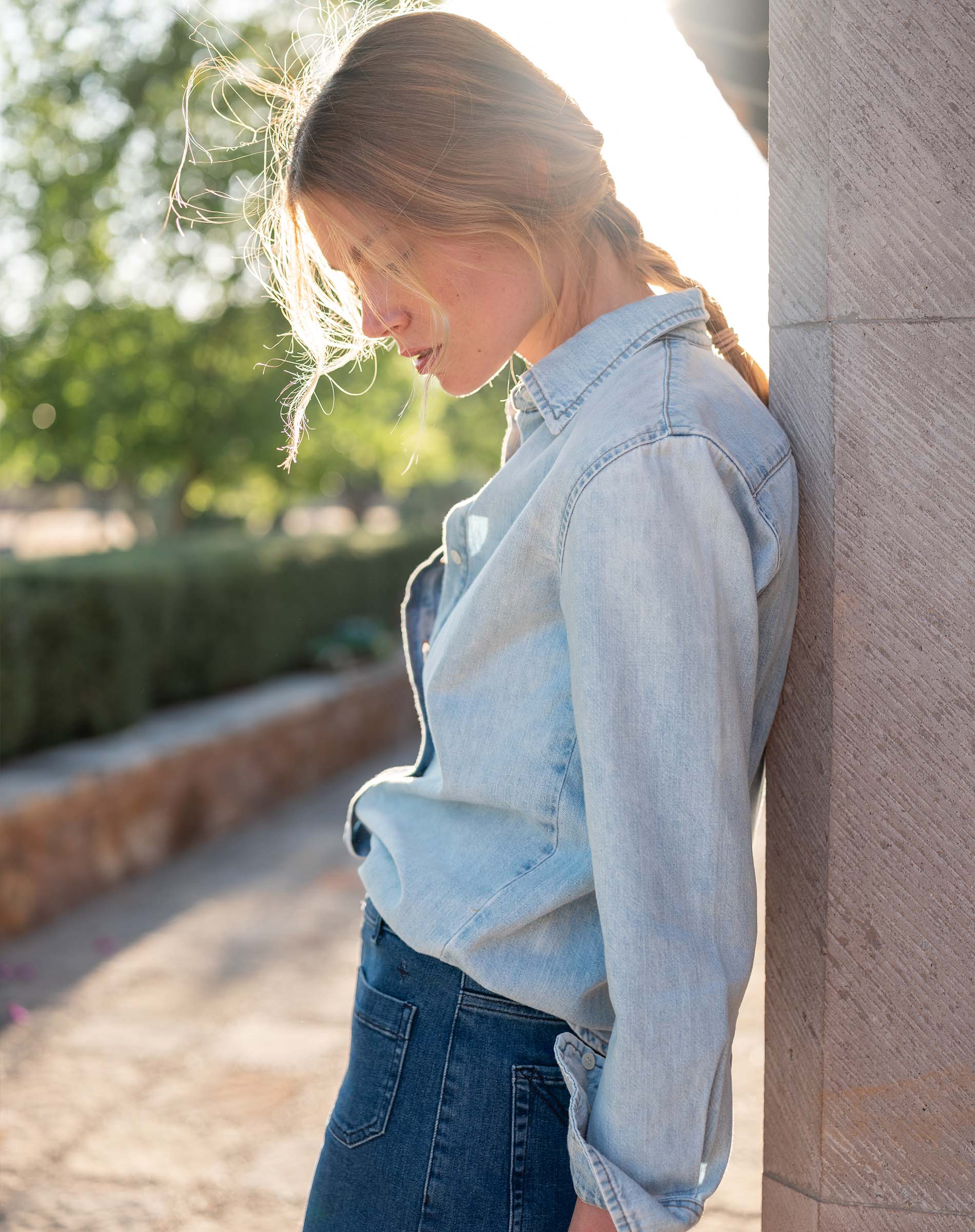 Woman wearing a light blue denim jacket and jeans leaning against a wall outdoors.