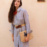 Woman wearing a striped dress with a belt, holding a woven bag against a beige wall.