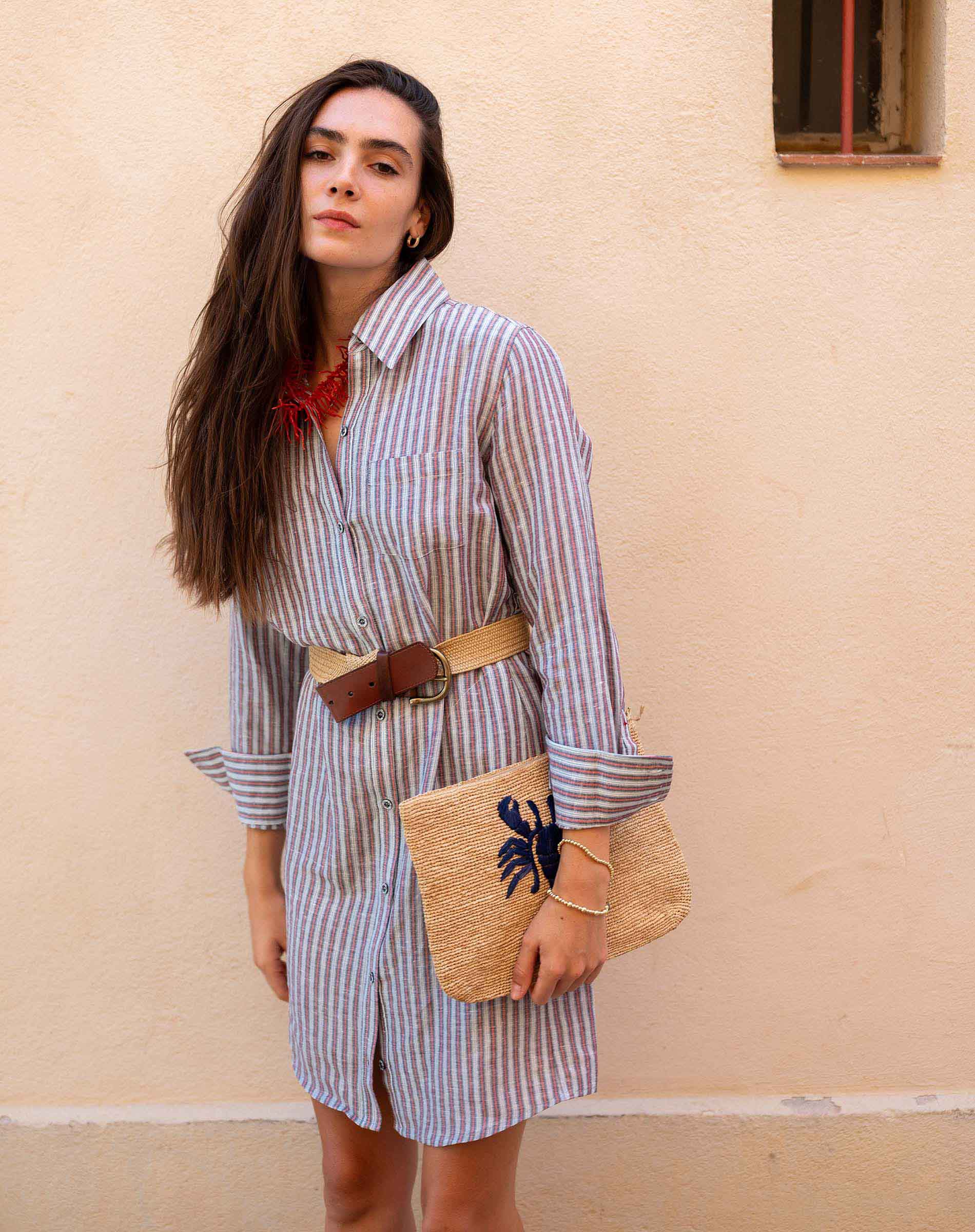 Woman wearing a striped dress with a belt, holding a woven bag against a beige wall.