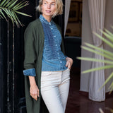 Model wearing an olive green kimono wrap over a blue textured button-down shirt and white trousers.