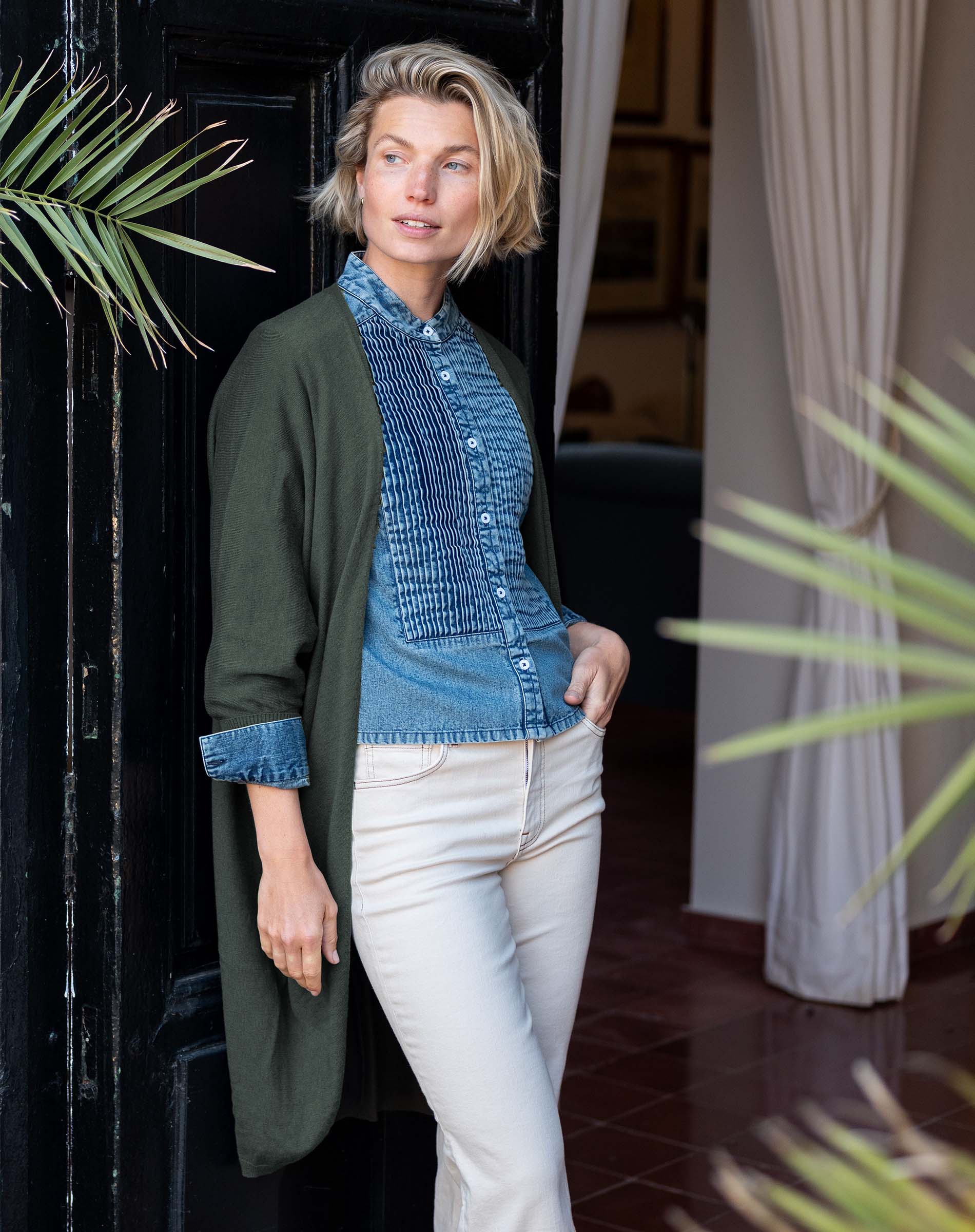 Model wearing an olive green kimono wrap over a blue textured button-down shirt and white trousers.
