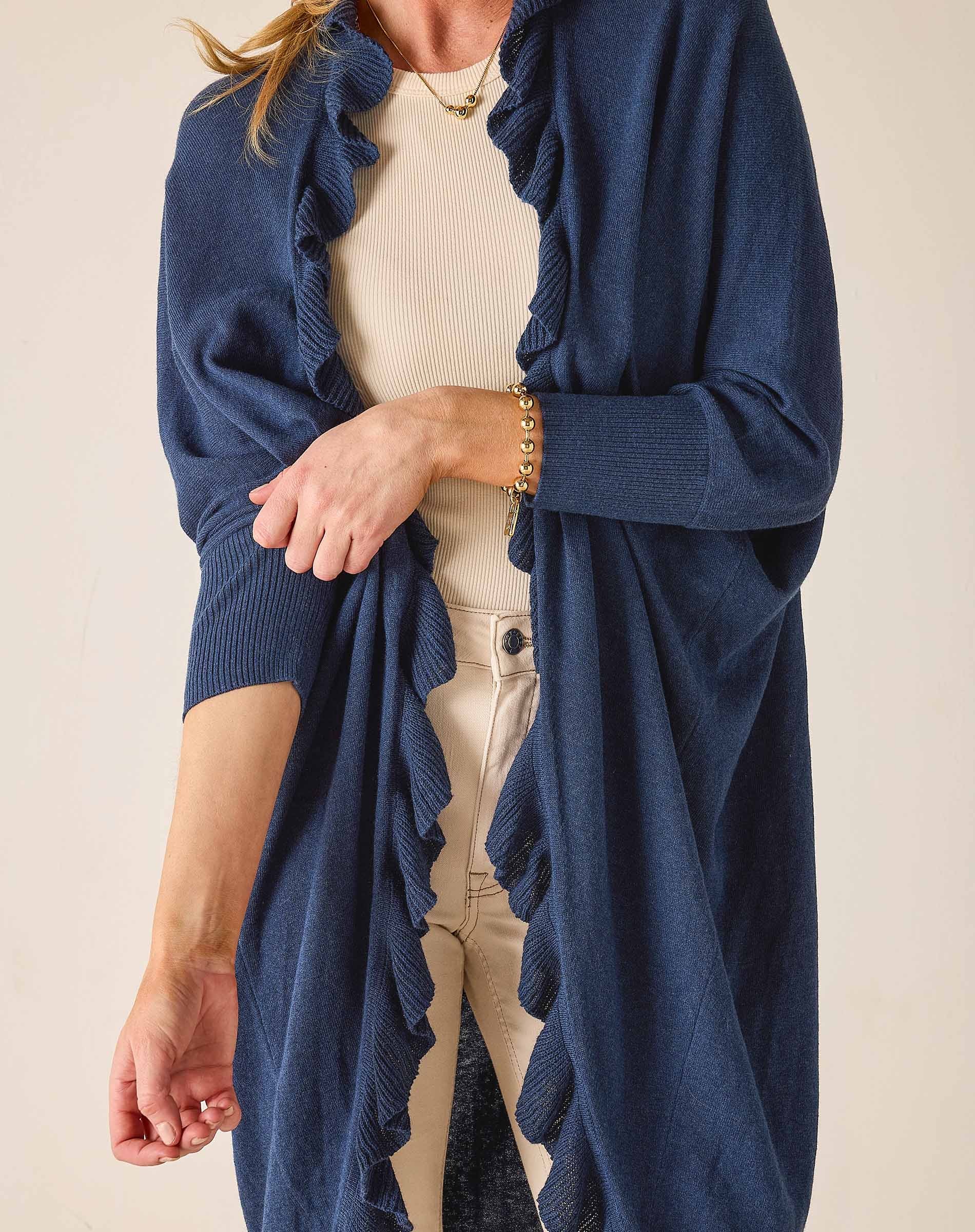 Person wearing a blue cardigan over a beige top and pants on a plain background