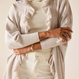Person wearing a beige cardigan over a white top with ruffled details, on a plain background.