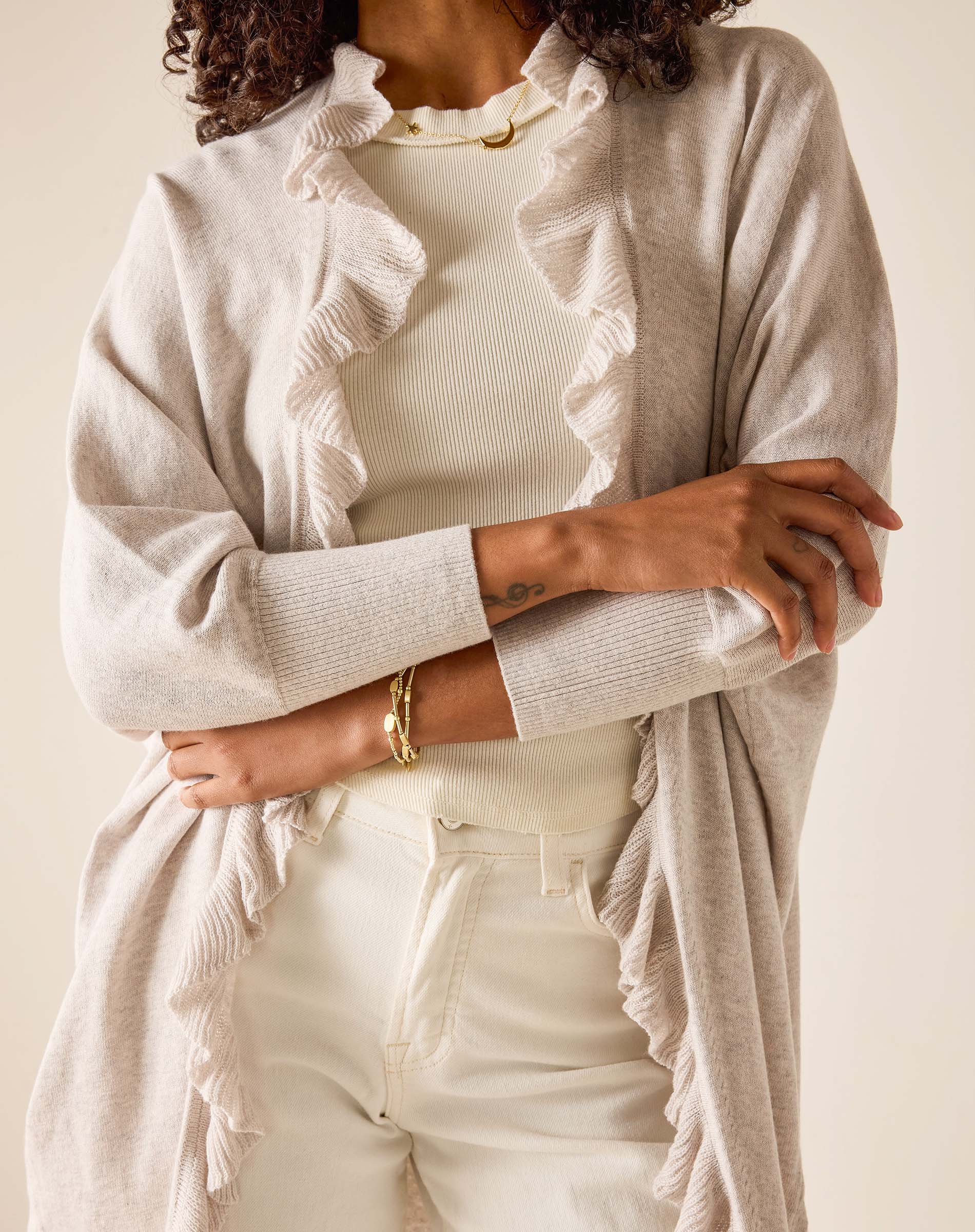 Person wearing a beige cardigan over a white top with ruffled details, on a plain background.