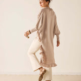 Woman wearing a beige long shirt and white pants against a plain background