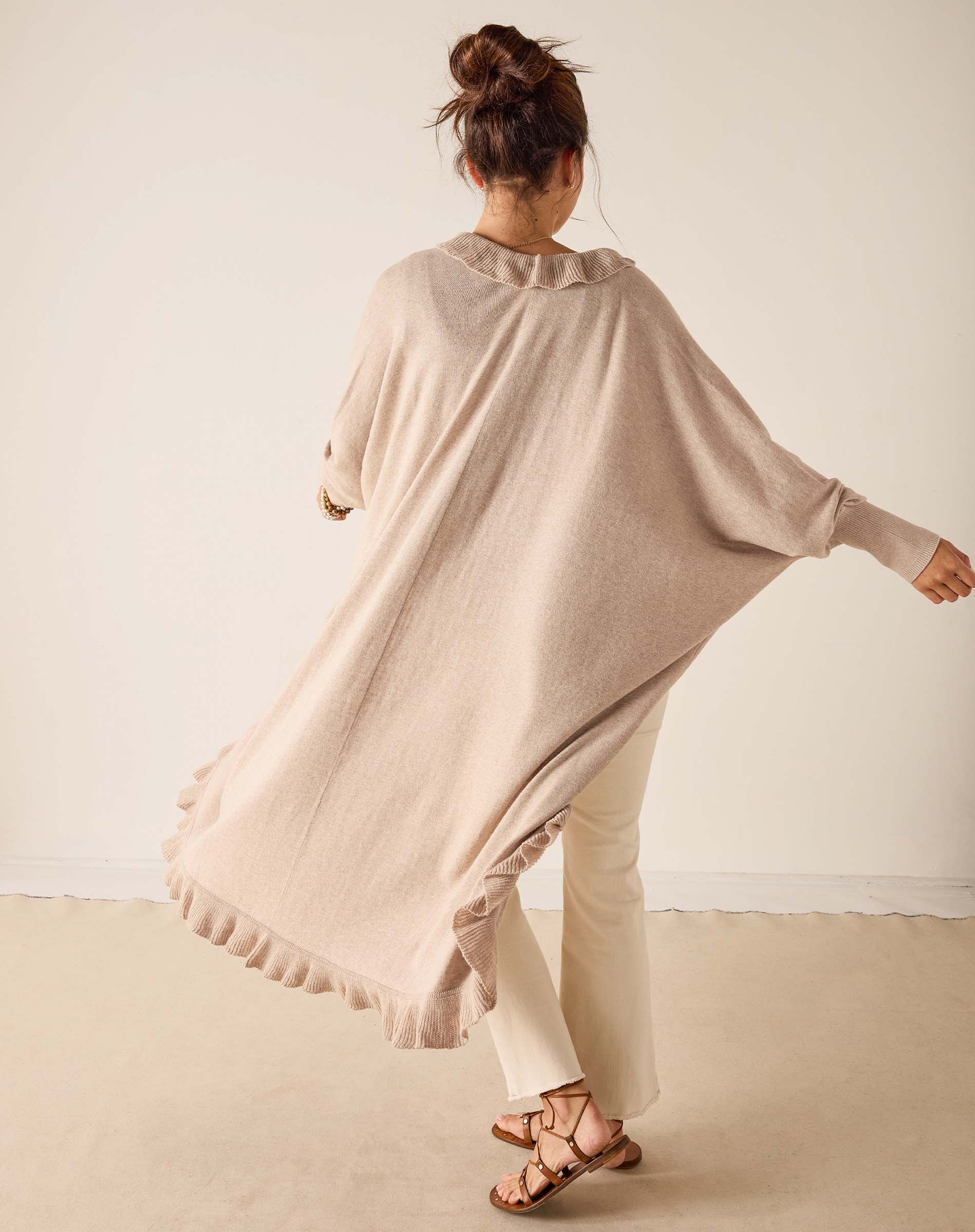 Woman wearing a beige poncho and white pants against a plain background