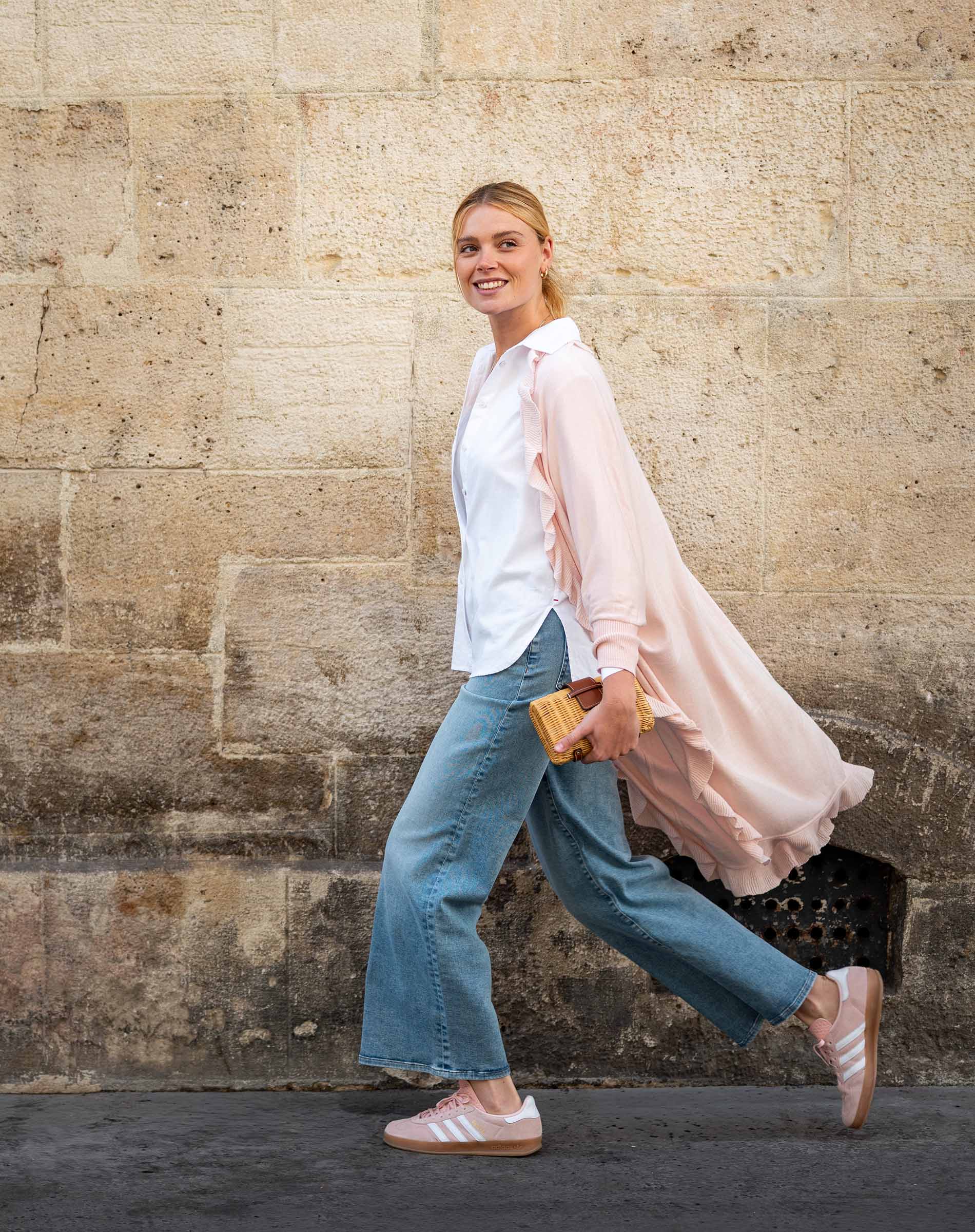 Woman walking against a stone wall wearing a white shirt, pink cardigan, blue jeans, and pink shoes.