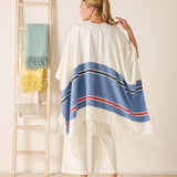 Woman wearing a white poncho with blue and red stripes, standing in front of a ladder with colorful cloths.